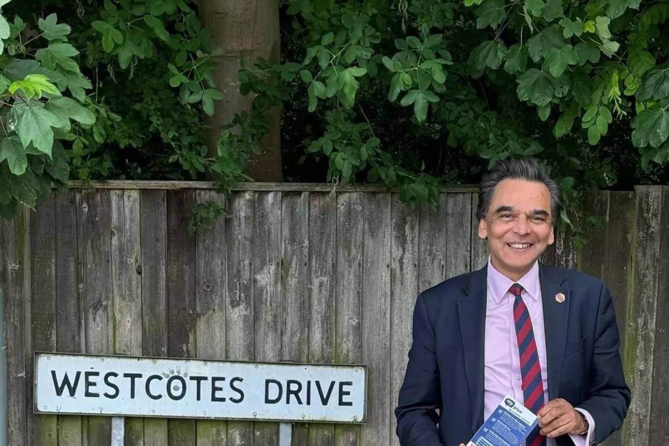 Photograph of Max Chauhan. He is smiling towards the camera, holding a leaflet, and standing in front of the Westcotes Drive sign and a wooden fence.