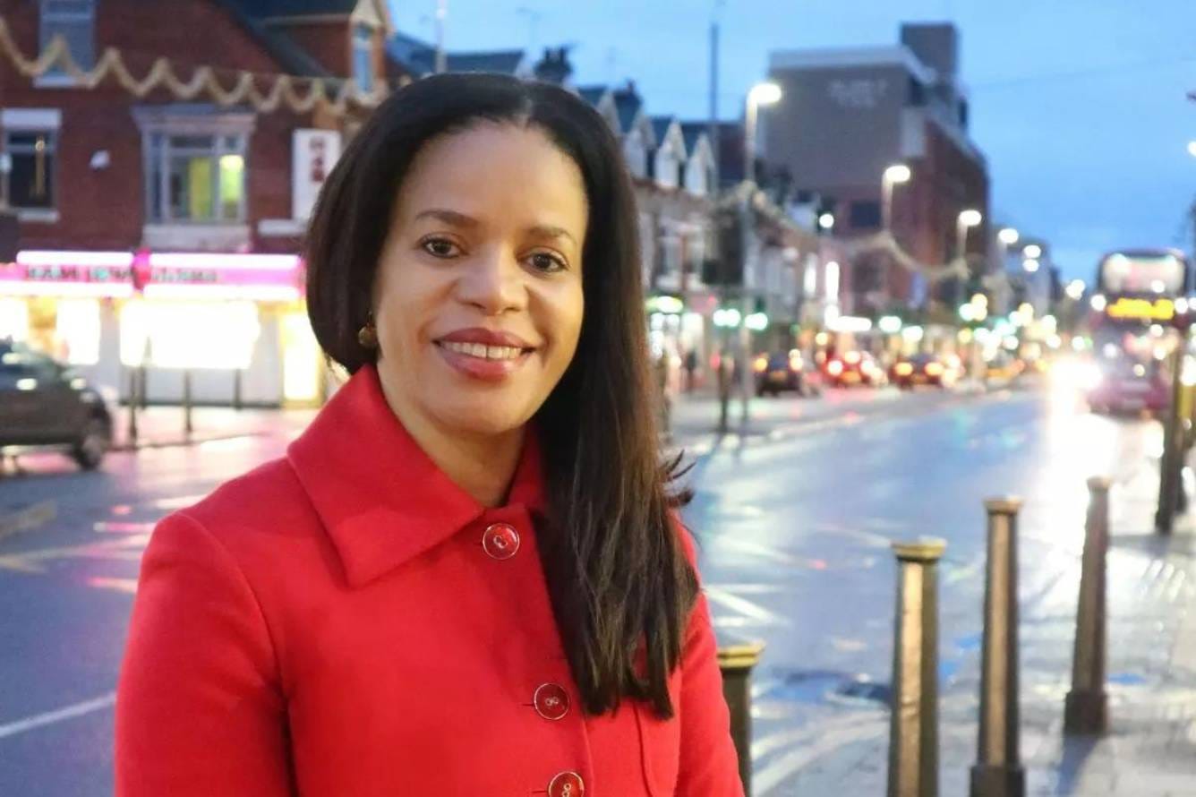 Photograph of Claudia Webbe, smiling towards the camera. She is wearing a red coat and standing on Belgrave Road in the evening. The street lights are on. There is a bus in the background.
