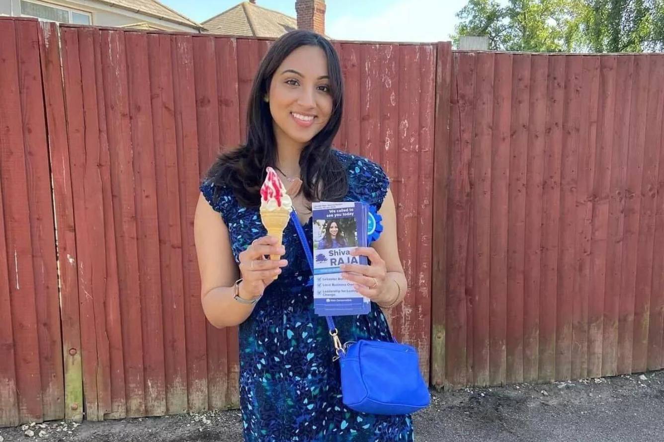 Photograph of Shivani Raja. She is smiling towards the camera, holding an ice cream and Conservative Party leaflet. There is a fence in the background. She is dressed in blue, the Conservative Party's colour.