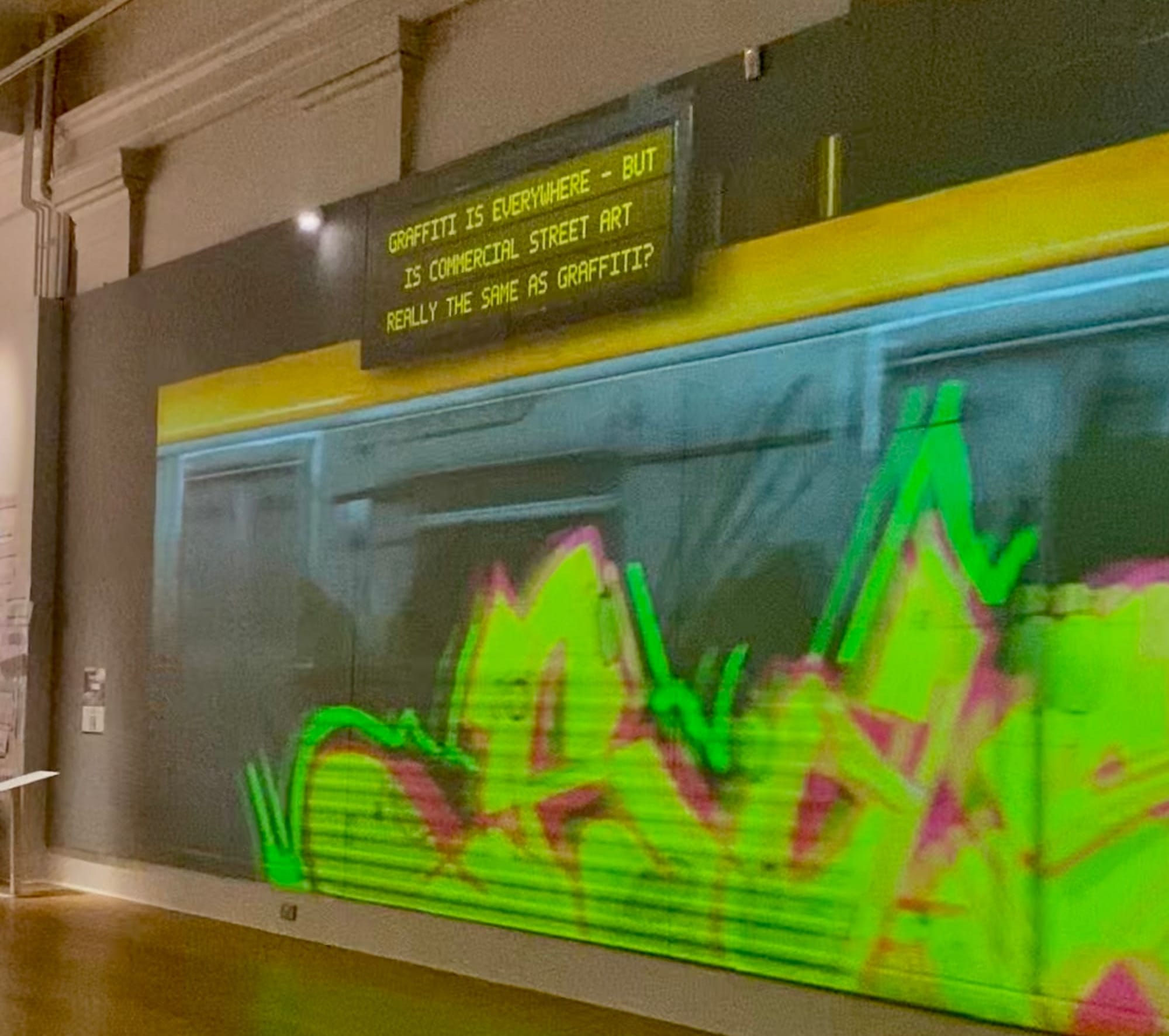 A street art subway train with a sign above it which reads 'Graffiti is everywhere - but is commercial street art really the same as graffiti?'