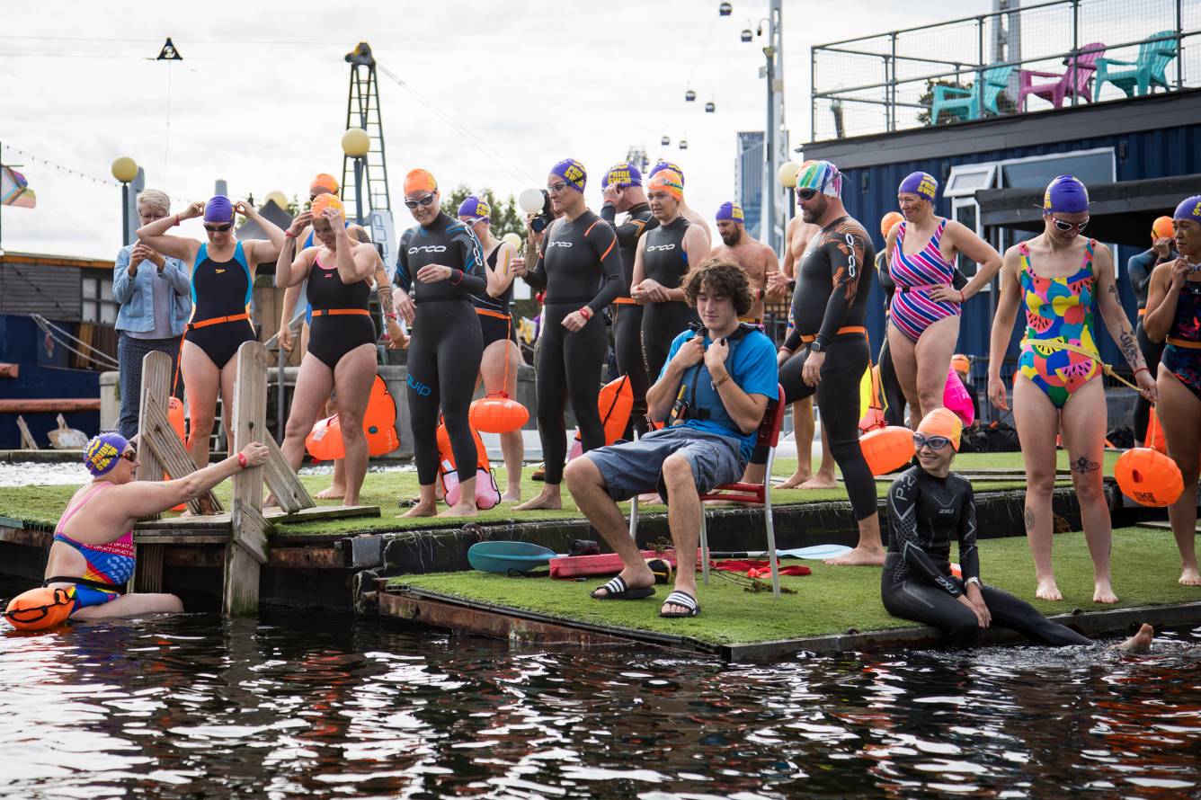 Several people gather around the edge of a river. Some are in the water, some are standing. They are all in swimming costumes.